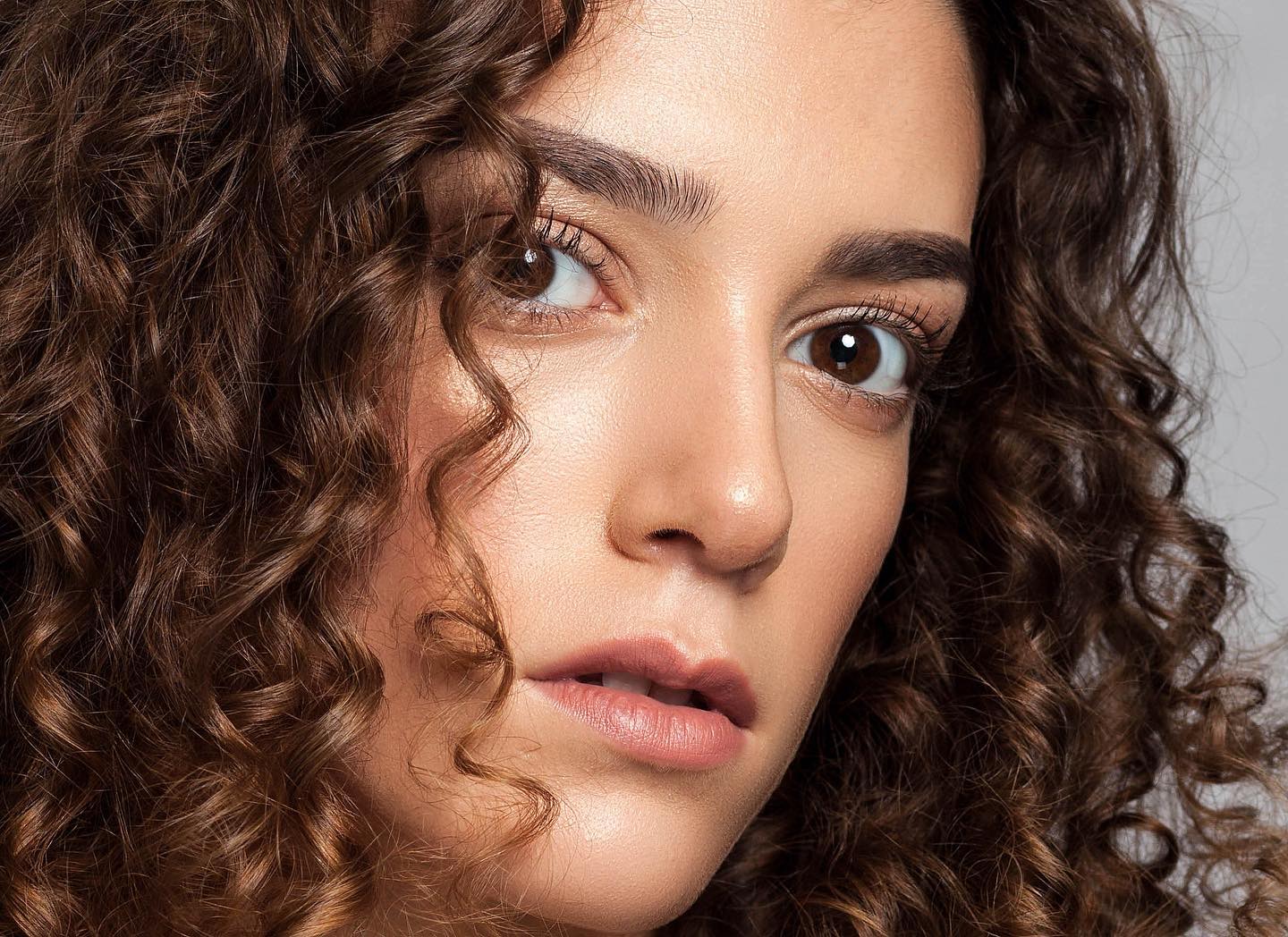 A brunette woman with curly hair gazes intently, her soft features highlighted by warm lighting in a close-up portrait.