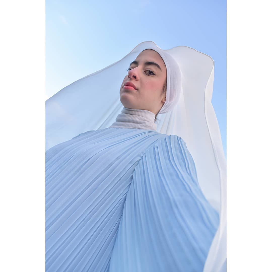 A woman in hijab poses in a light blue dress against the sky, blending fashion with serenity.
