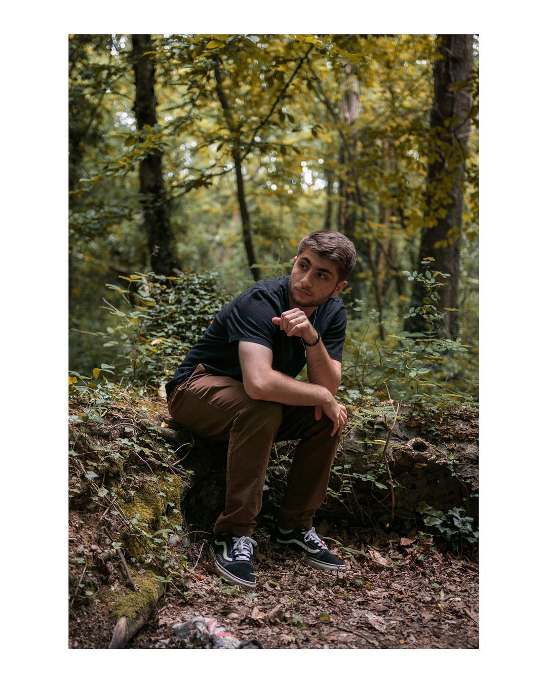 A brunette man sits contemplatively in a forest setting, captured in a medium shot.