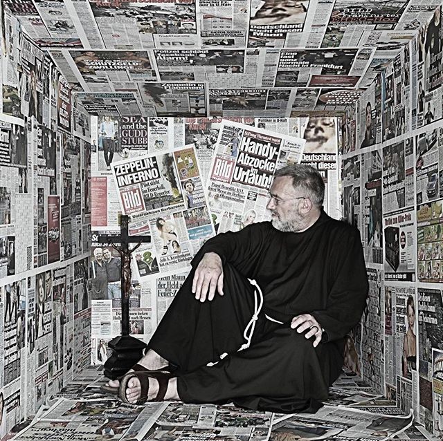 A man dressed as a friar sits surrounded by newspapers, conveying a sense of isolation and information overload.