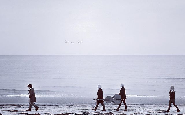 Four women walk along a beach, one carrying a cello, in a monochrome scene of ocean and sky.