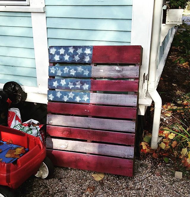 A rustic American flag made from a wooden pallet leans against a light blue house.