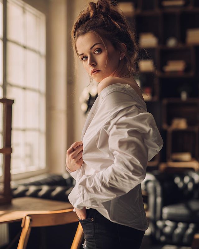 A woman in a white shirt poses in a room with natural light and bookshelves, creating a dreamy and intimate portrait.
