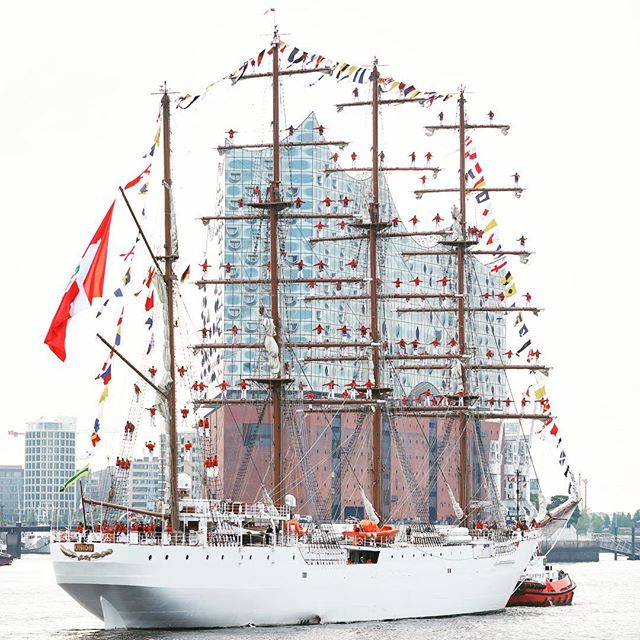 A tall ship with sails is decorated with flags and people in front of a modern glass building on the water.