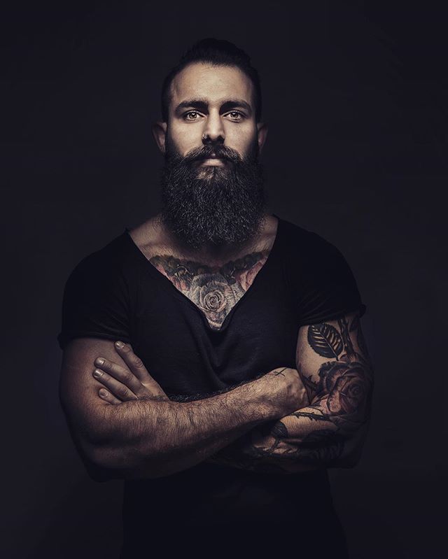A man with tattoos and a beard is seen from the waist up with his arms crossed.