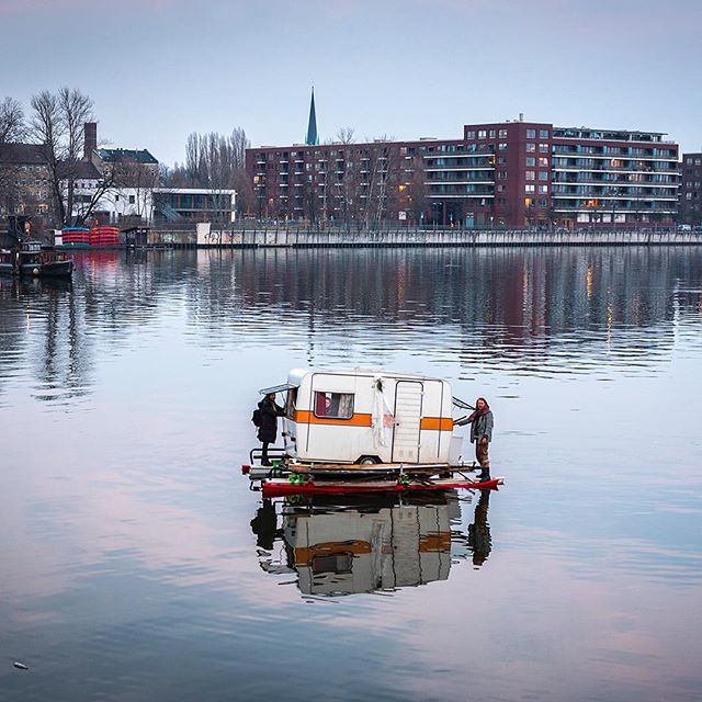 Two people on a camper raft float down a river with urban buildings in the background, reflecting in the water.