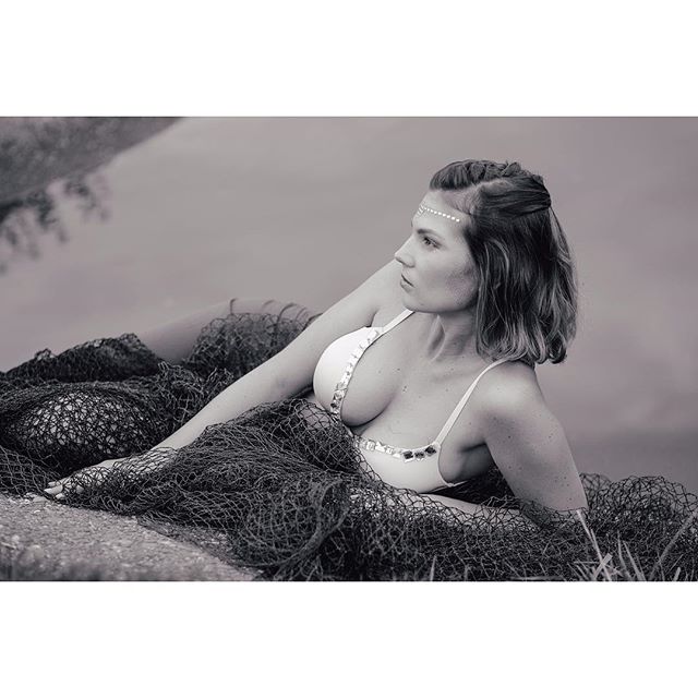 A woman in a bikini poses in a fishing net near the water's edge in a monochrome setting.
