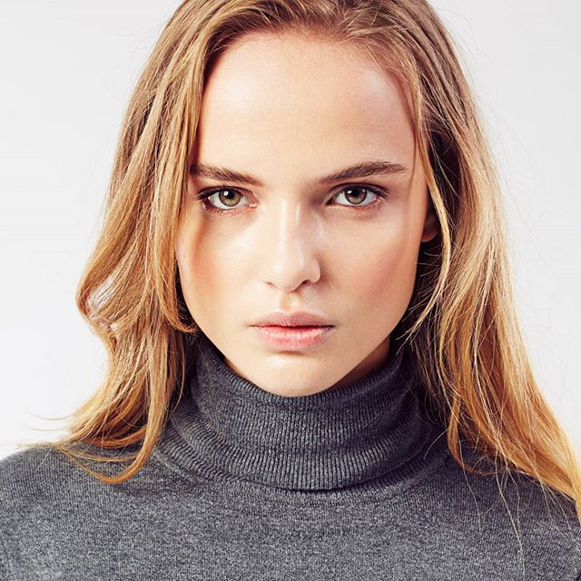 A woman with blonde hair and a gray turtleneck stares intensely in a modern portrait.