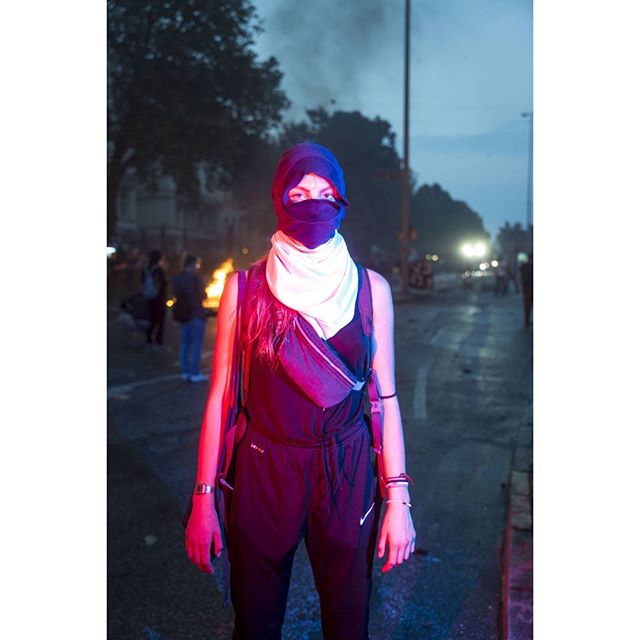 A masked woman stands defiantly amidst a street protest, bathed in an eerie mix of blue and red light.