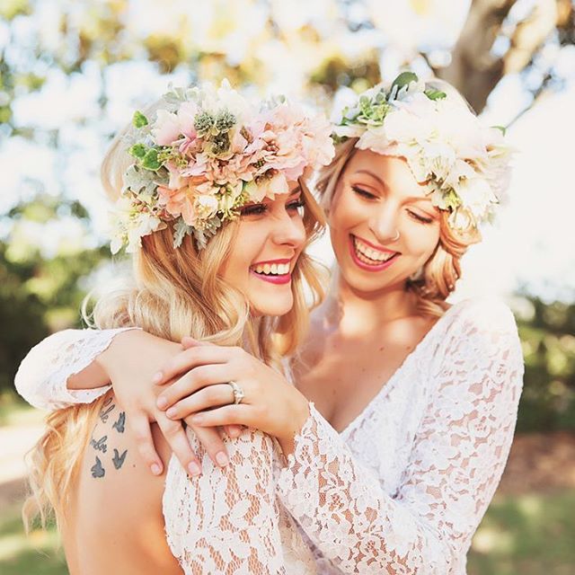 Two cheerful women with flower crowns embrace, wearing lace dresses, perfect for a bridal or romantic themed visual.