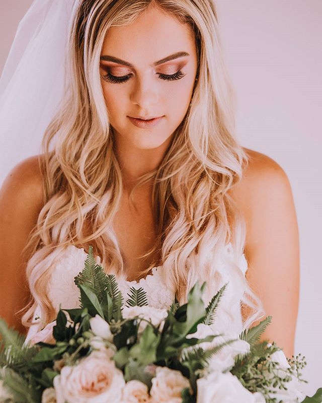 A beautiful bride with blonde hair holds a wedding bouquet in a softly lit studio setting, looking down with a dreamy expression.