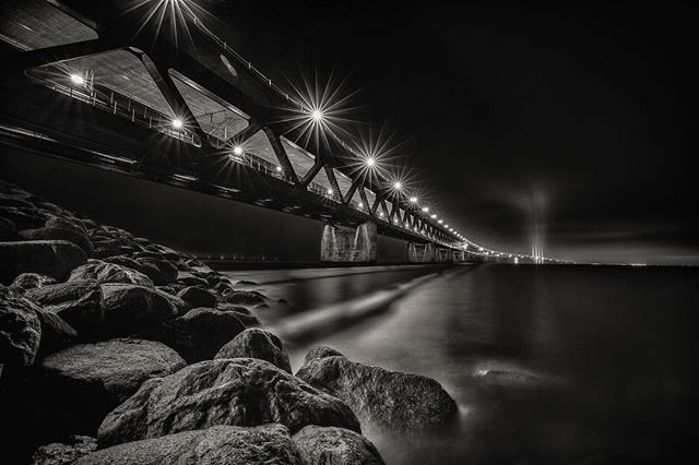 A black and white long exposure shows a bridge illuminated at night over calm water near rocks.