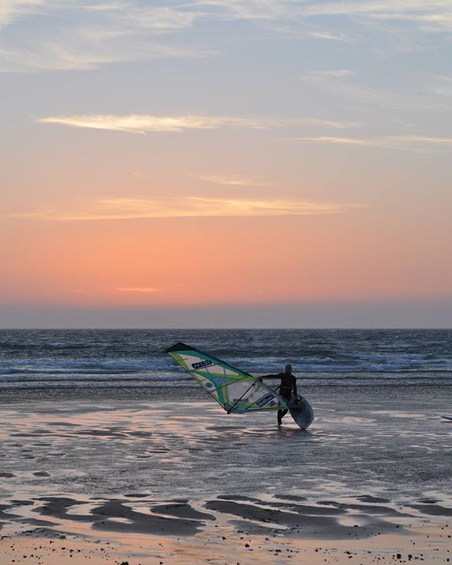 A windsurfer walks on the beach at sunset with a sailboard, enjoying a calm and relaxing ocean view.