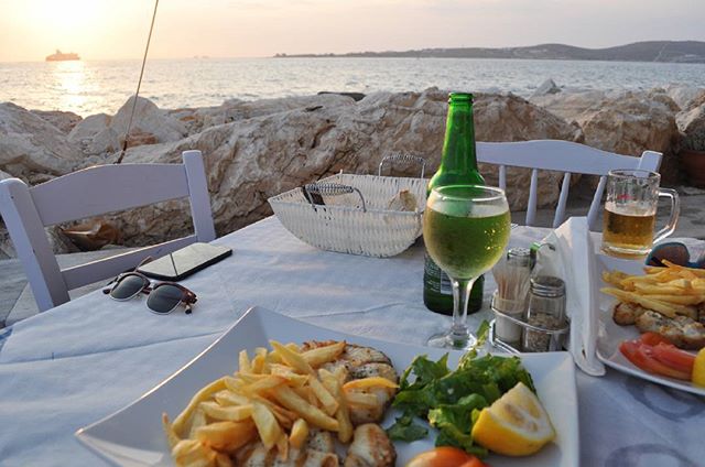 A table with food, drinks, and accessories at a restaurant overlooking the ocean at sunset, for a relaxed dining experience.