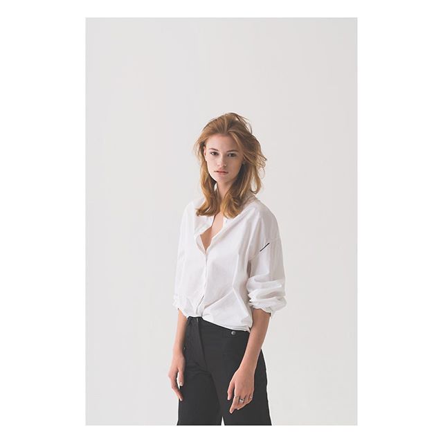 A poised woman with red hair models a white shirt and black pants against a clean white backdrop.