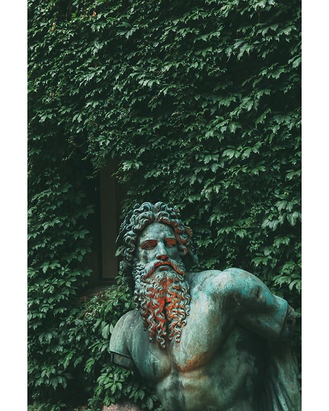 A weathered statue of a bearded man stands against a backdrop of lush green foliage.