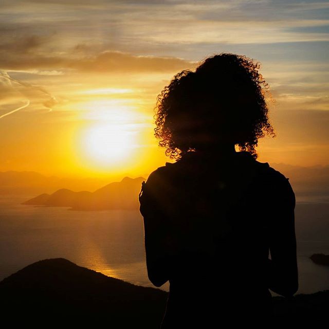 A woman's silhouette looks out at the ocean and a vibrant sunset over mountains.