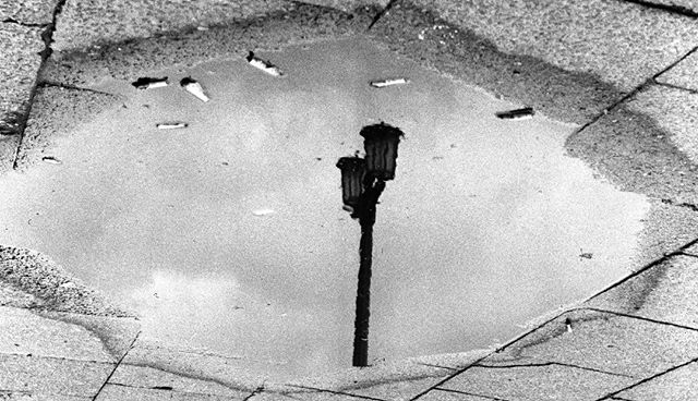 A street lamp is reflected in a puddle on a city sidewalk, surrounded by cigarette butts.
