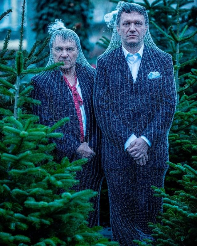 Two men stand among Christmas trees wearing netting over suits in a humorous image.