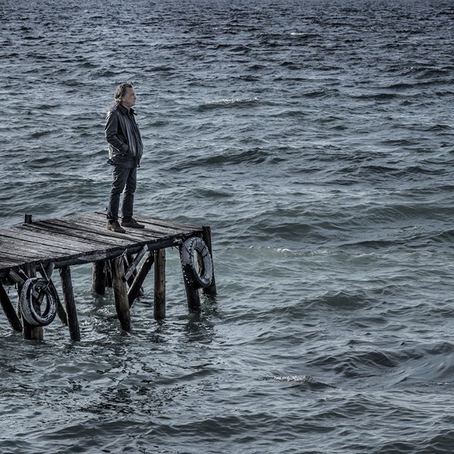 A man stands on a weathered pier looking out at the rough sea in a contemplative moment.