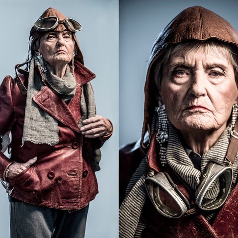 An elderly woman with a determined expression poses in vintage aviator attire for an editorial portrait.