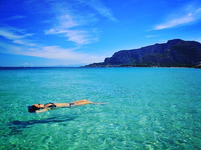 A woman relaxes floating on her back in clear turquoise ocean water on a bright, sunny day.