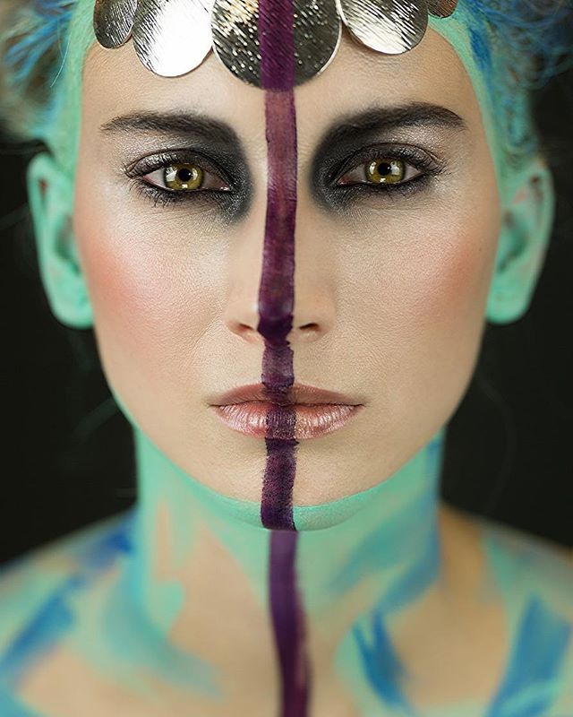 A woman with striking makeup and a teal face is seen in an editorial-style close-up.