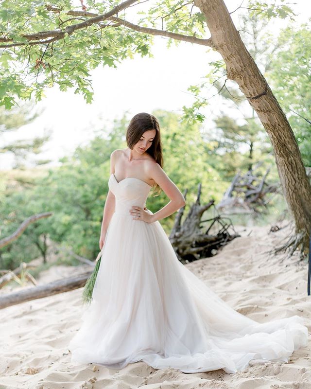 A bride in a flowing white wedding dress poses gracefully on a sandy beach with a forest backdrop, creating a dreamy and romantic scene.