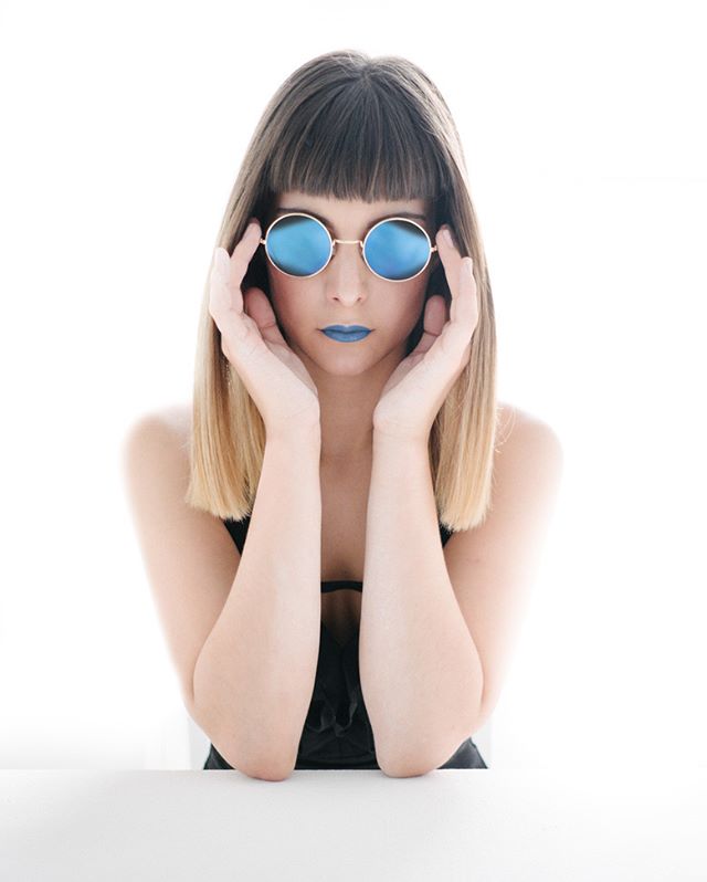 A woman with blue lipstick and round blue sunglasses poses in a brightly lit studio.