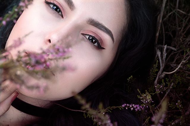 A brunette woman with pink makeup is lying in nature amongst flowers.