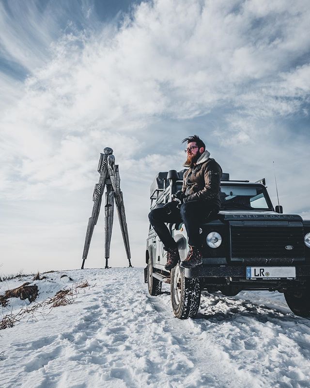 A bearded man sits on a Land Rover in a snowy landscape, next to a large sculpture, enjoying the winter scenery.