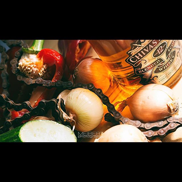 A bottle of whisky is surrounded by onions, pepper and a chain, creating a moody and evocative still life composition.