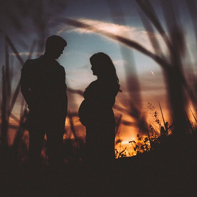 Silhouette of a pregnant woman and her partner embrace at sunset in a grassy field, celebrating their soon-to-be family.