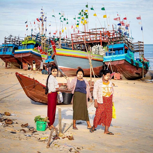 Three women stand on a sandy beach with colorful fishing boats lined up in the background under a cloudy sky.