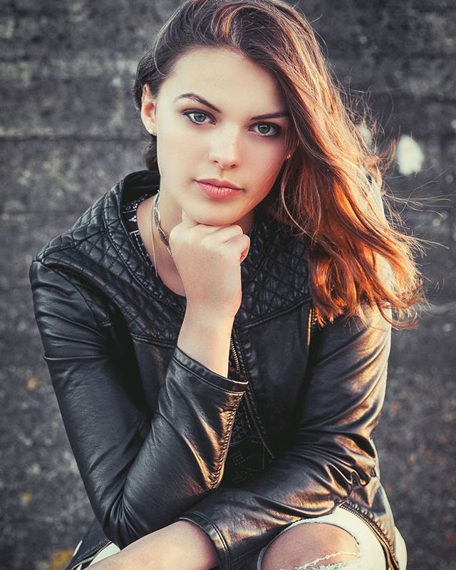 A young woman with auburn hair poses confidently in a black leather jacket against an urban backdrop.