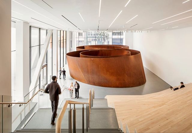 An indoor art gallery showcases a large, rust-colored sculpture with visitors exploring the space.