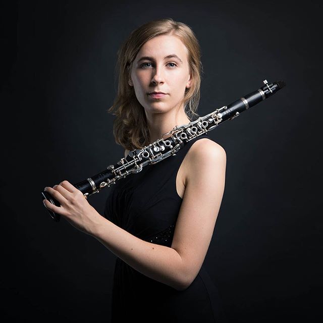 A blonde woman in a black dress poses with a clarinet against a dark background.