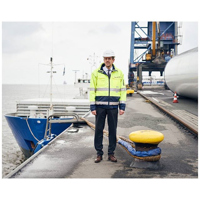 A man in workwear stands at a port with a ship and crane in the background.