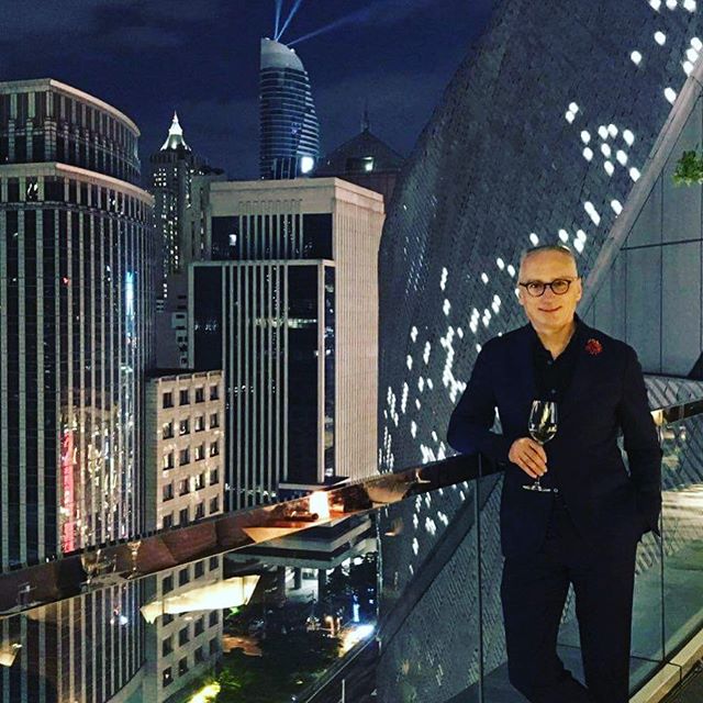A stylish man relaxes on a balcony overlooking a glittering cityscape at night.