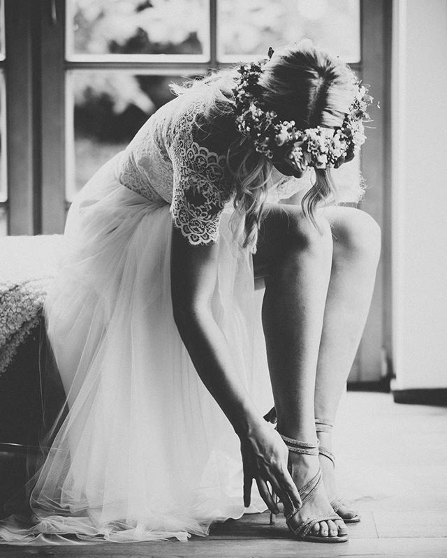 A bride in a flower crown fastens her shoes, bathed in soft light, in an elegant, timeless image.