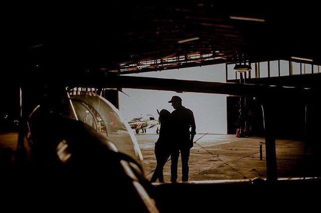 Silhouetted couple embracing in an airplane hangar with a small aircraft behind them.