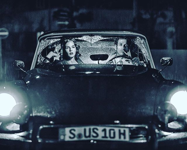 Stylish couple rides in a car on a rainy night, evoking a film noir mood.