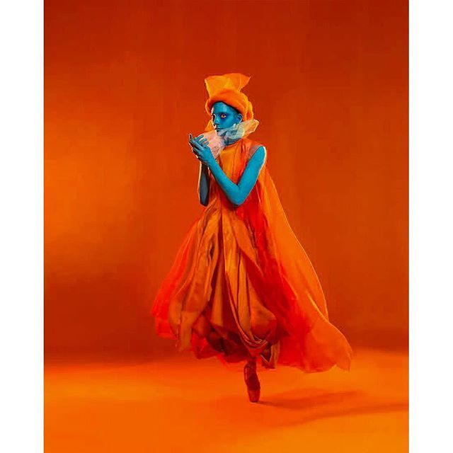 A blue-skinned ballerina in an orange dress performs a surreal dance in a vibrant studio setting.