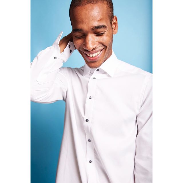 A smiling man in a white dress shirt poses against a blue backdrop, showcasing modern, minimalist style.