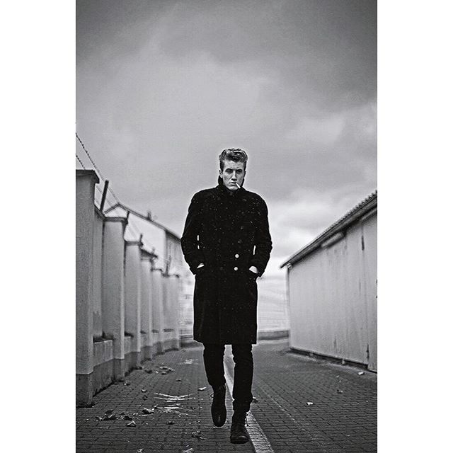 A man in a coat walks down a street with a cigarette in a black and white editorial style photograph.
