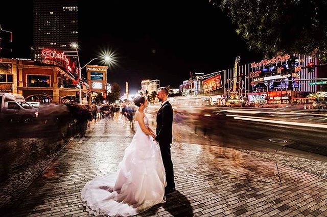 A bride and groom stand on a lit city street at night, traffic streaks by, and many people move in the background.