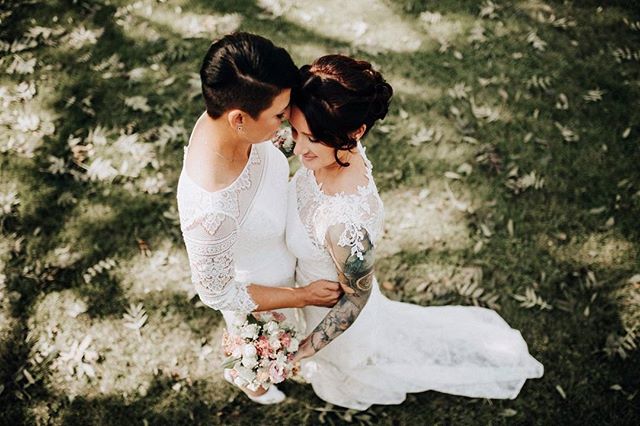 Two brides in lace wedding dresses hold a bouquet at their outdoor wedding.