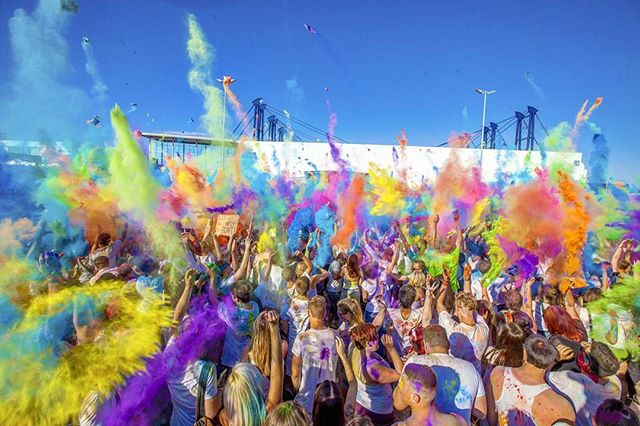 A large crowd celebrates Holi, throwing colorful powder into the air at an outdoor festival.