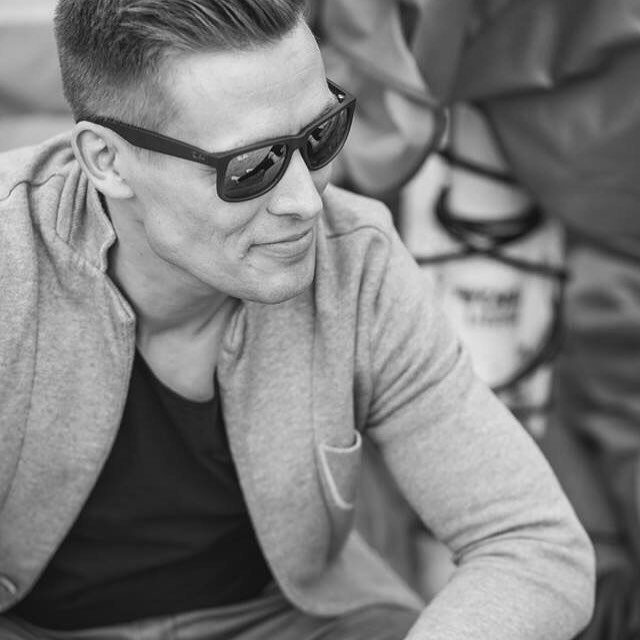 A man in sunglasses and a jacket poses in a black and white outdoor shot.