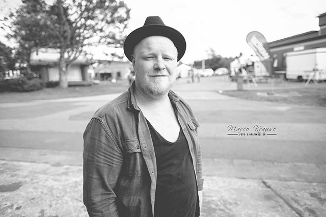 A man in a hat and open shirt stands outside in a black and white portrait.